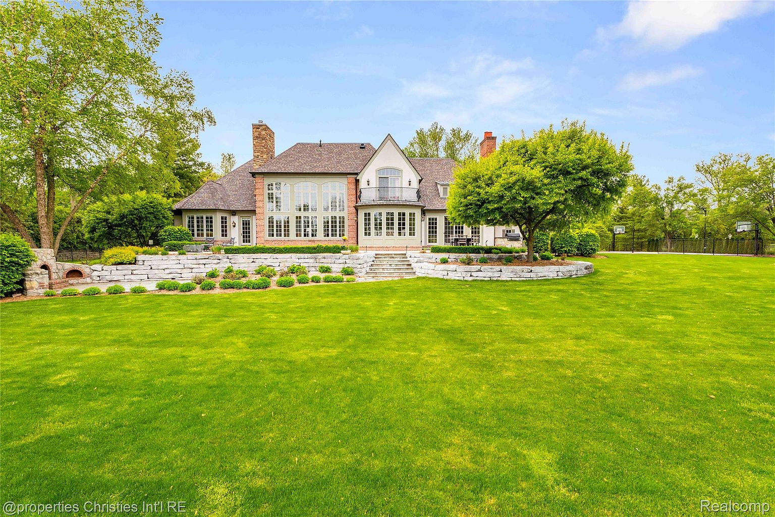 This image showcases the rear exterior of a large, luxurious home with a well-manicured lawn. The house features multiple windows, a balcony, and a stone retaining wall with landscaping. A basketball court is visible in the background, suggesting ample outdoor recreational space.