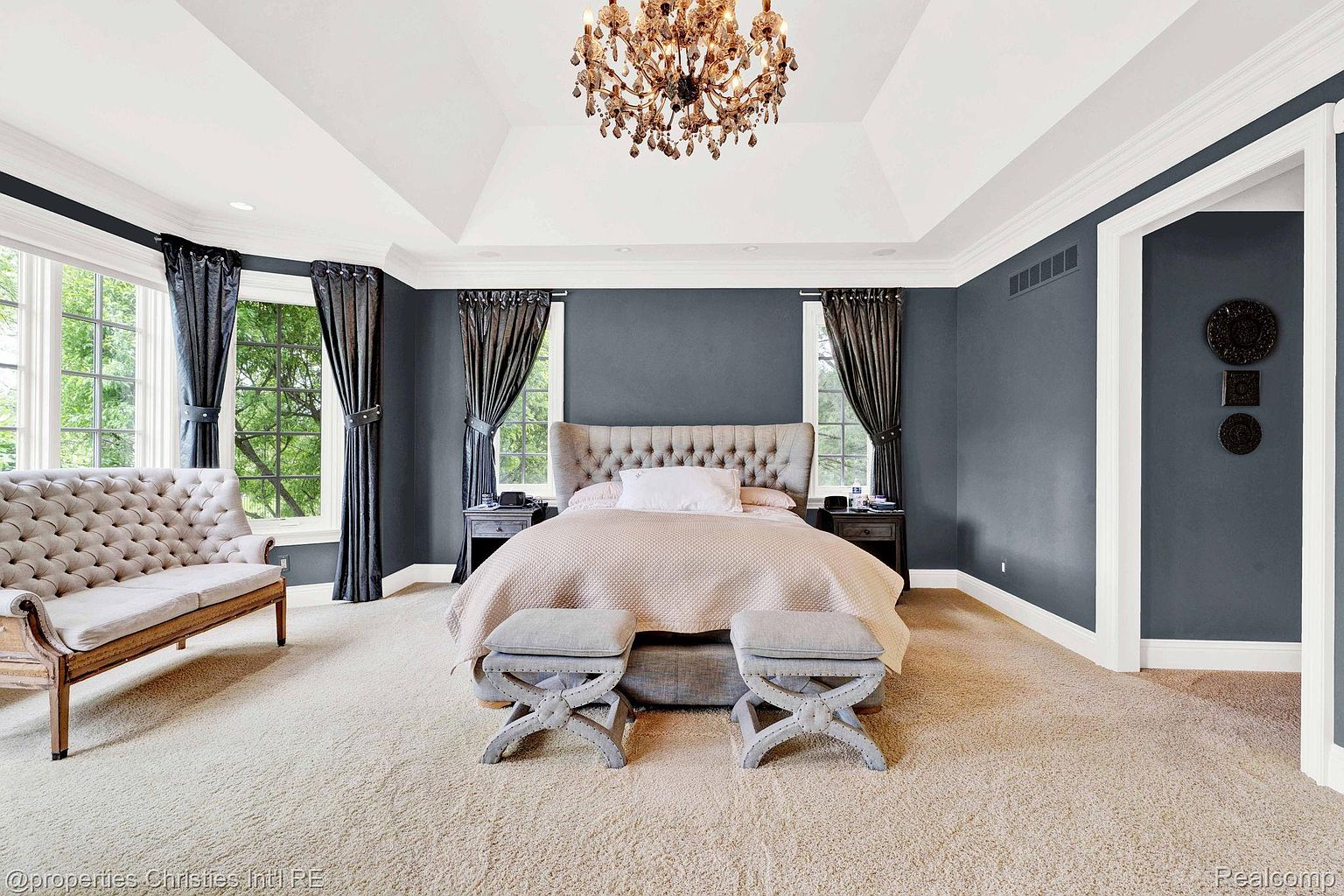 This is a luxurious primary bedroom featuring a tufted headboard bed with neutral bedding, flanked by matching nightstands and windows dressed with elegant dark curtains. The room is illuminated by a grand chandelier, and the walls are painted in a sophisticated dark gray, creating a serene and upscale ambiance. Two upholstered benches are placed at the foot of the bed.