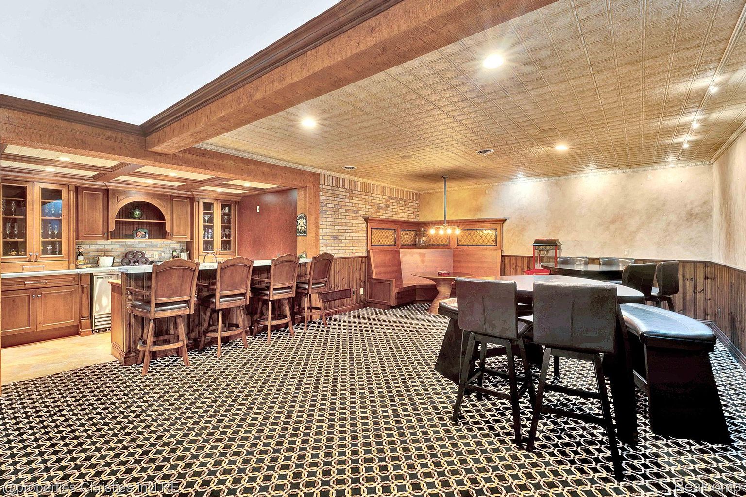 This is a finished basement featuring a bar area with wooden cabinetry and bar stools, a dining area with booth seating, and a patterned carpet. The ceiling has a textured, possibly metallic, tile design, and the overall style is reminiscent of a classic pub or lounge. The image is taken from a low angle, emphasizing the depth of the space.