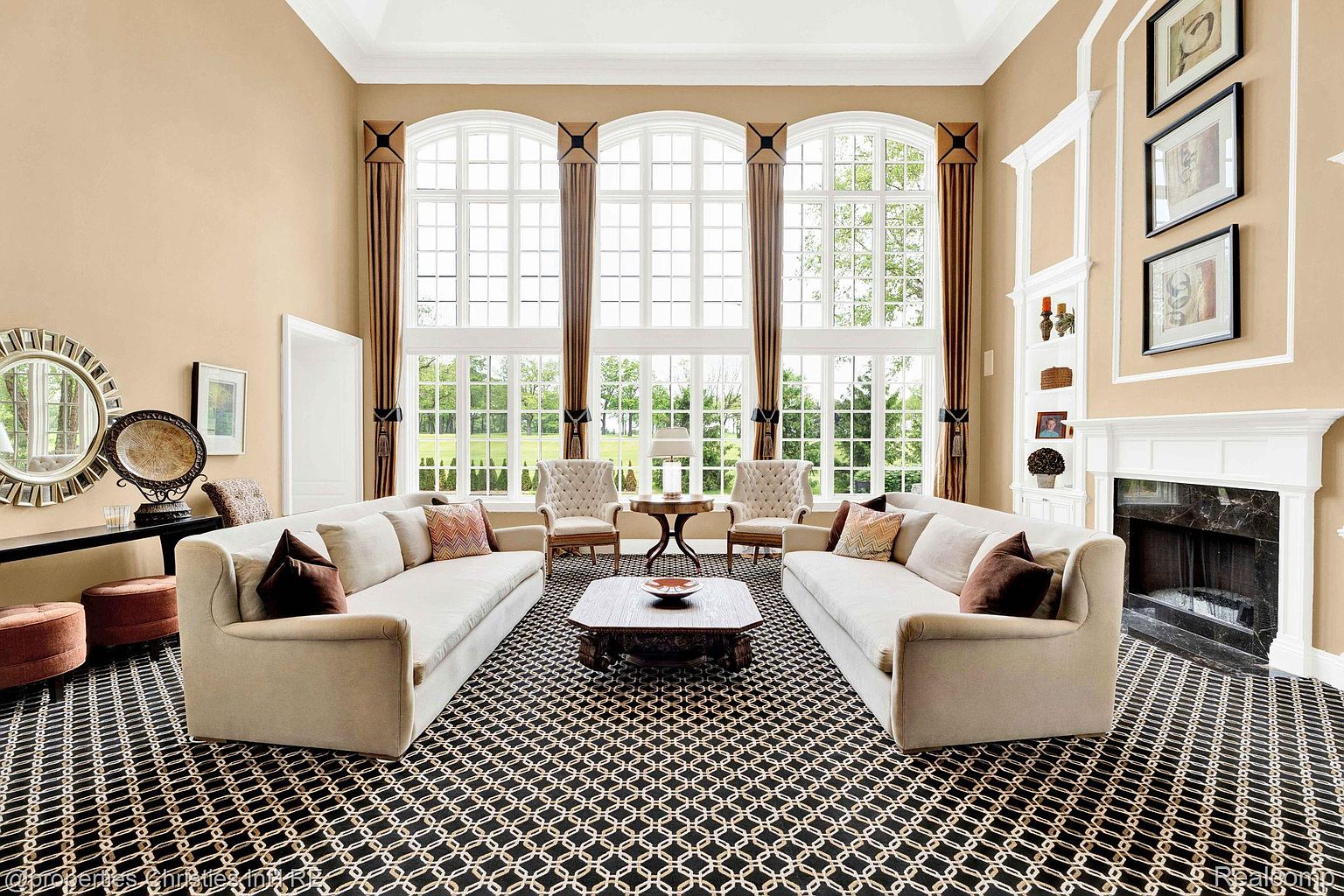 This is a spacious and elegant living room featuring two large sofas facing each other, centered around a decorative coffee table. The room is bathed in natural light from three large arched windows, complemented by floor-to-ceiling drapes. A black and white patterned rug covers the floor, and a fireplace with a white mantel adds a focal point to the room.