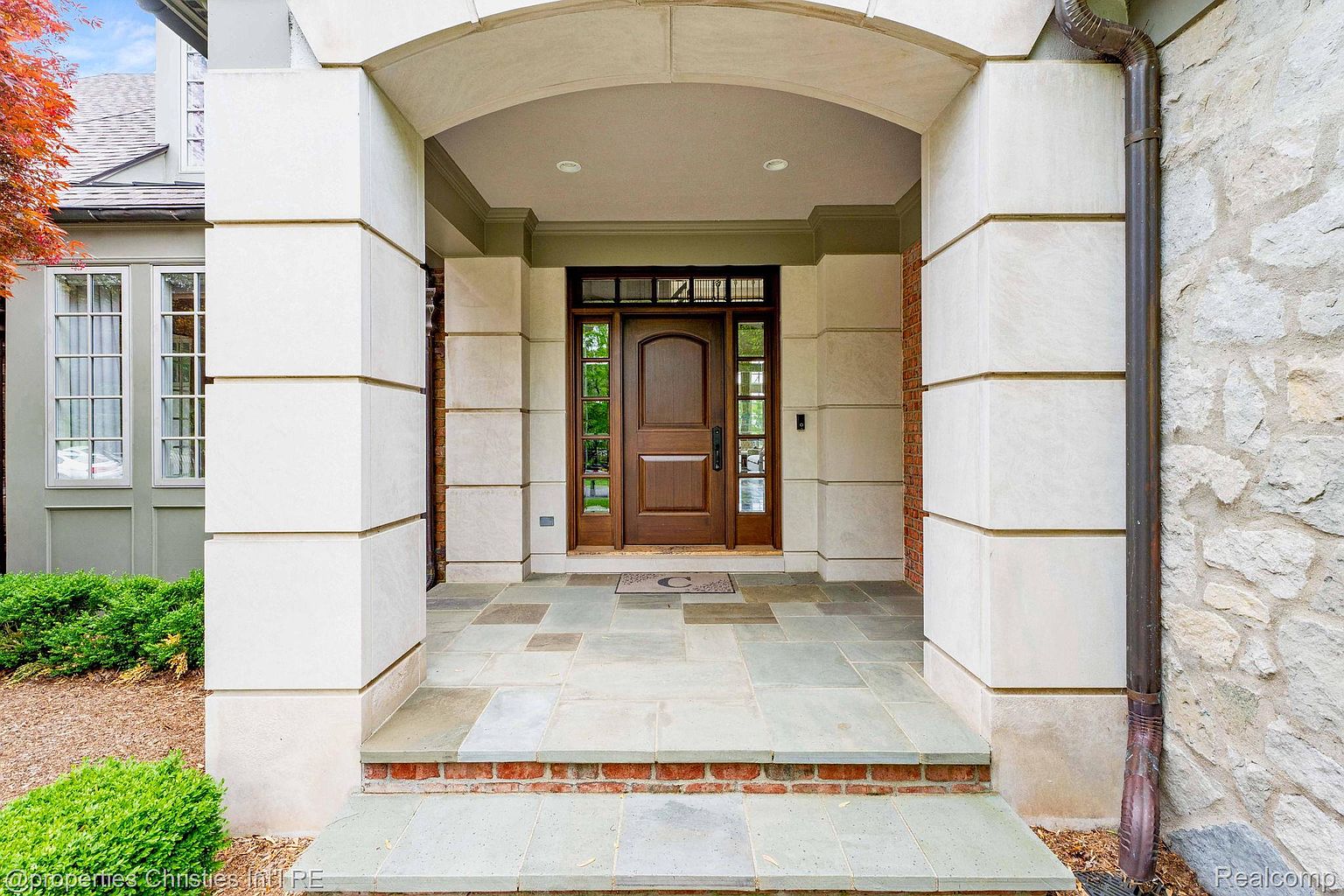 This image showcases the grand entryway of a home, featuring a solid wood front door with glass panels, flanked by substantial stone pillars and an arched stone lintel. The entryway is paved with large, square stone tiles, and a small brick step adds a touch of traditional charm. The overall impression is one of stately elegance and welcoming warmth.