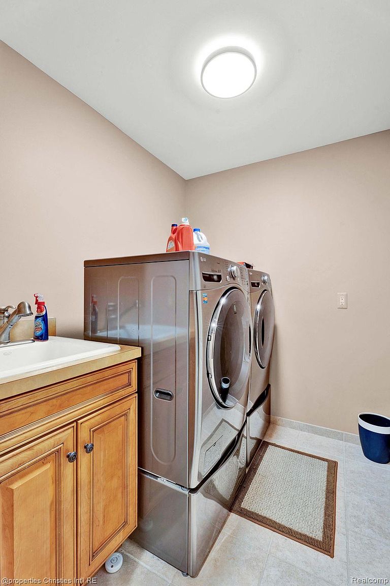 This is a well-organized laundry room featuring a front-loading washer and dryer set on pedestals. A sink with a wooden cabinet is located to the left, providing a convenient space for handwashing. The room is brightly lit with a flush mount ceiling light and has a neutral color scheme, creating a clean and functional space.