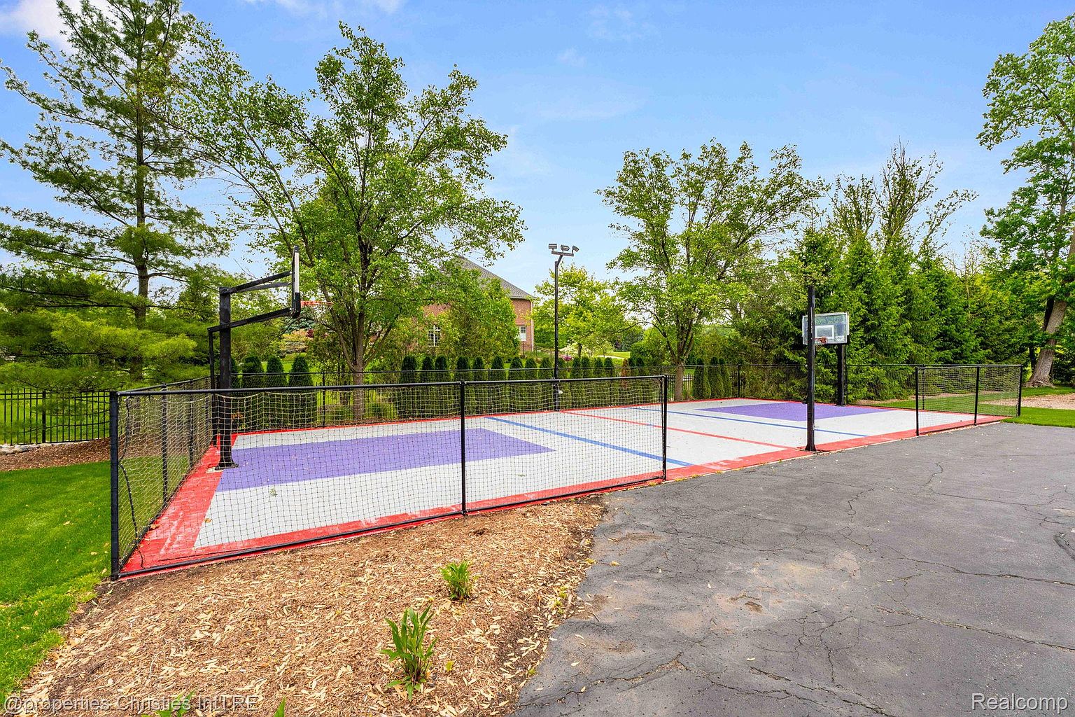 This image showcases a well-maintained outdoor basketball court, enclosed by a black fence and netting. The court features a colorful playing surface with red, white, and purple sections, and is surrounded by landscaping and a paved area. Mature trees provide shade and enhance the property's appeal.