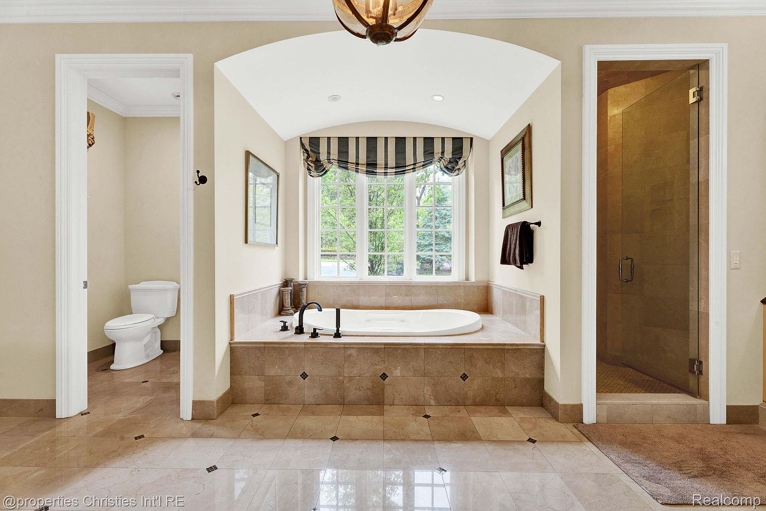 This is a luxurious primary bathroom featuring a large soaking tub centered beneath a window with a striped valance. The bathroom has tiled floors and walls, with a separate toilet room and glass-enclosed shower visible on either side. The overall impression is one of elegance and relaxation.