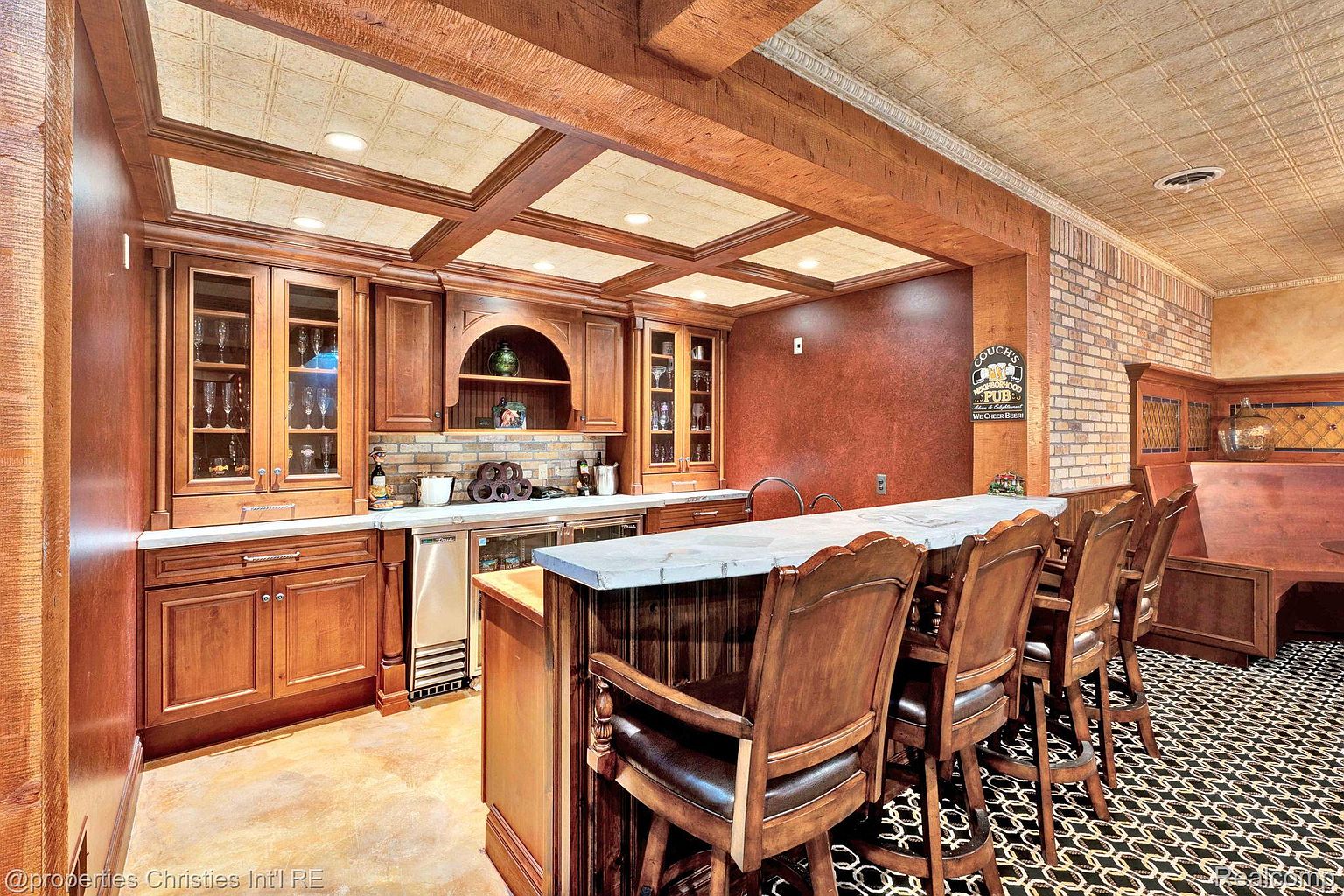This image showcases a well-appointed basement bar area, featuring rich wood cabinetry, a marble countertop, and stylish bar stools. The coffered ceiling with decorative tiles adds a touch of elegance, while the brick accent wall and patterned flooring contribute to the room's character. The space is designed for entertaining and relaxation.