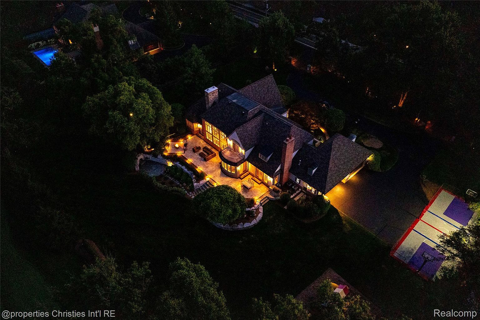 This aerial view showcases a luxurious home at night, highlighting its well-lit patio, manicured lawn, and adjacent sports court. The house features a complex roofline, multiple chimneys, and warm interior lighting visible through large windows, creating an inviting and upscale impression. A swimming pool is also visible in the distance.