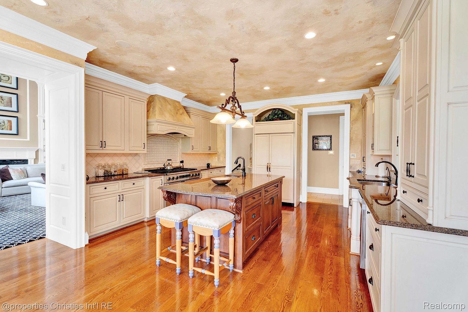 This is a well-lit kitchen featuring custom cabinetry, granite countertops, and a central island with seating. The kitchen includes high-end appliances and a decorative range hood. The hardwood floors add warmth to the space, creating an inviting and luxurious atmosphere.
