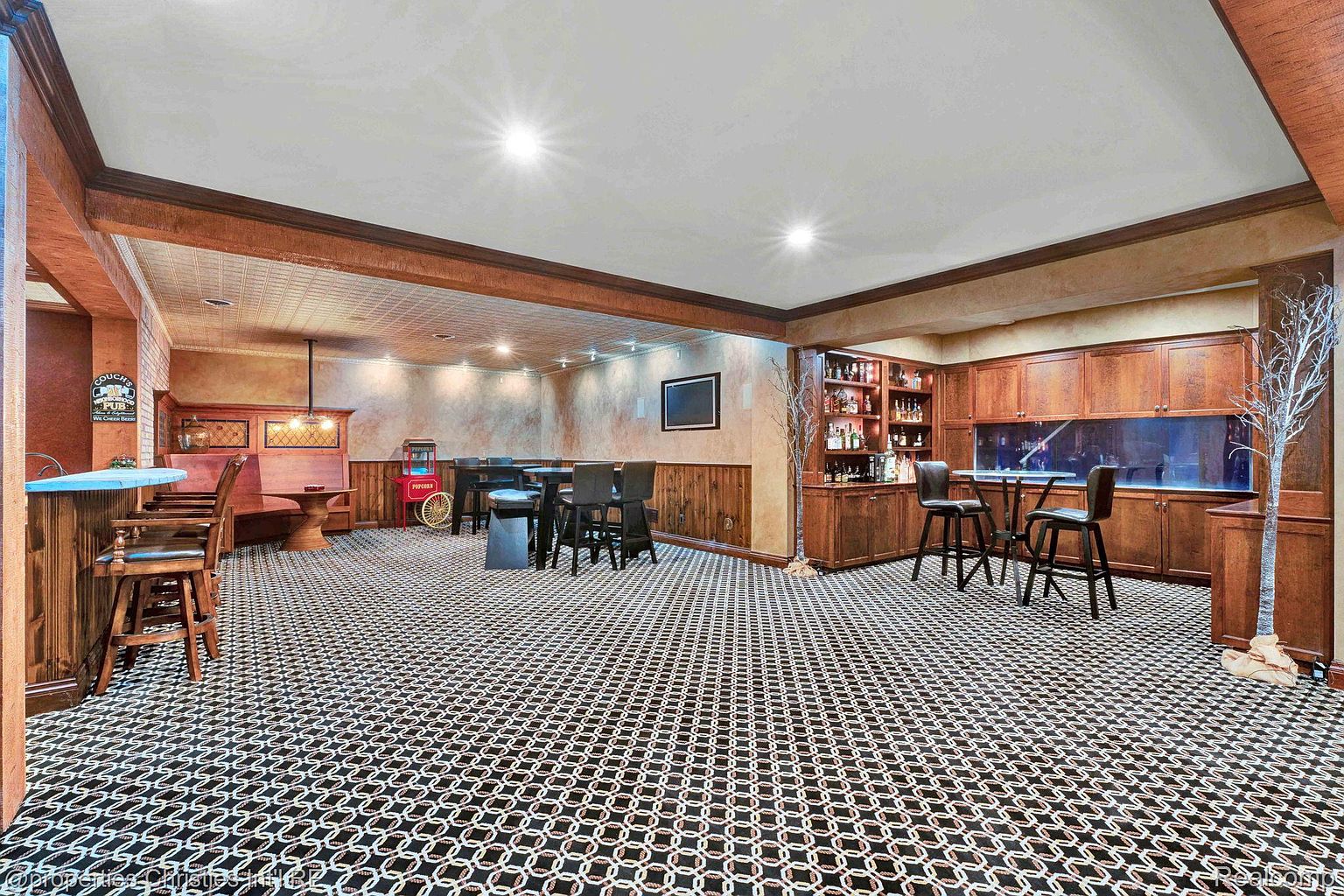 This is an interior shot of a finished basement, designed as an entertainment space. The room features a bar area with seating, a dining table with chairs, and a built-in cabinet with a large fish tank. The patterned carpet and warm wood tones create a cozy and inviting atmosphere.