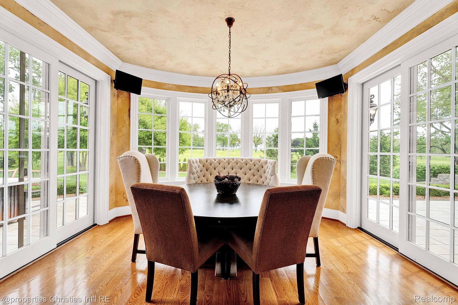 This is an interior shot of a dining room featuring a round table surrounded by upholstered chairs and a tufted bench. Large windows offer a view of the exterior landscape, and a decorative chandelier hangs above the table. The room is well-lit with natural light, creating a warm and inviting atmosphere.