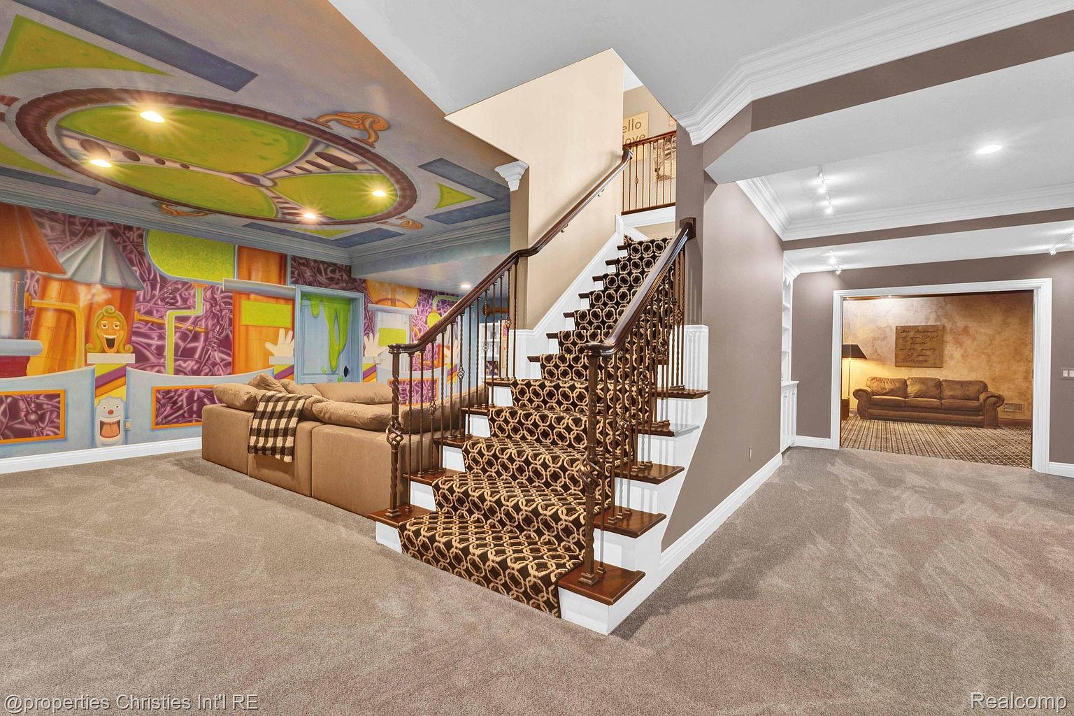 This is a spacious basement area featuring a whimsical mural on the wall and ceiling, a comfortable brown sectional sofa, and a staircase with patterned carpeting. The room is well-lit and appears to be designed as a recreational or entertainment space. The perspective is from the corner of the room, capturing both the living area and the staircase leading to another level.
