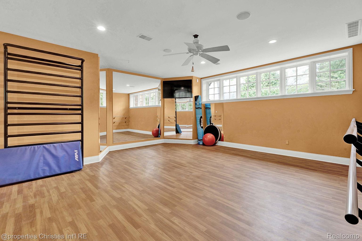 This is a spacious home gym featuring wood-look flooring and warm-toned walls. The room includes a wall-mounted ladder bar with a blue mat, a wall of mirrors, a flat-screen TV, and a window providing natural light. The overall impression is a clean and well-equipped space for exercise.