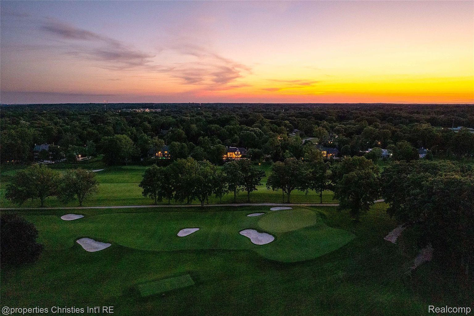 This aerial view showcases a lush golf course community at sunset. Several houses are nestled among the trees, with the vibrant colors of the sky reflecting on the manicured greens. The image conveys a sense of luxury, tranquility, and exclusivity, highlighting the desirable location and lifestyle.