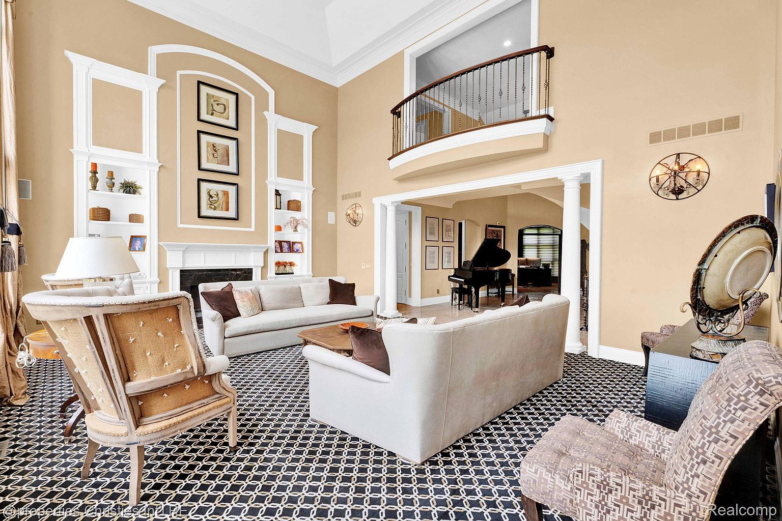 This is an interior shot of a luxurious living room featuring two sofas, an ornate armchair, and a patterned black and white rug. The room has high ceilings, built-in shelving flanking a fireplace, and a balcony overlooking the space. A grand piano is visible in an adjacent room, suggesting a sophisticated and elegant home.