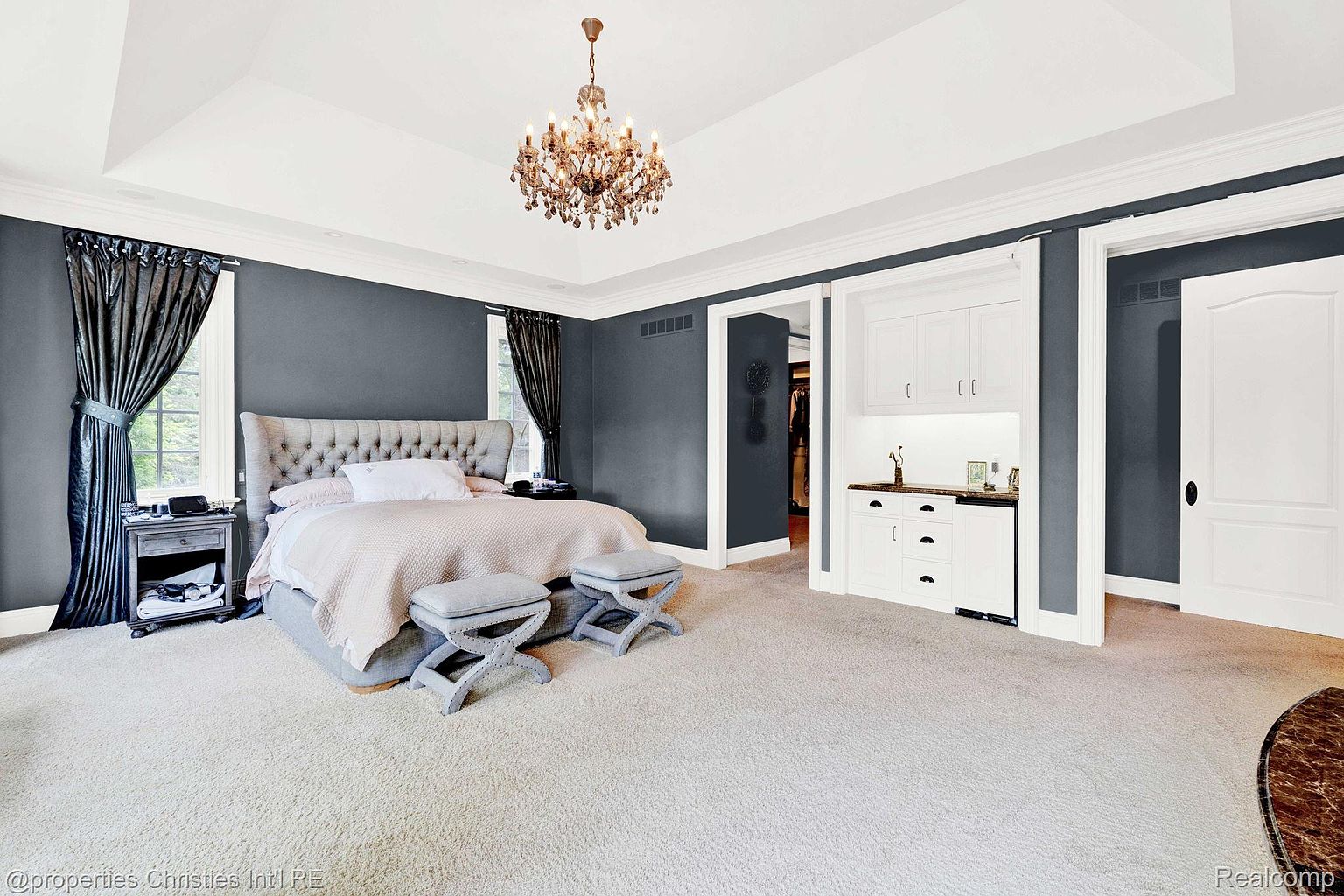 This is a primary bedroom featuring a large bed with a tufted headboard, two decorative ottomans, and a crystal chandelier. The walls are painted a dark gray, and the room has white trim and a light-colored carpet. There is a built-in bar area and a closet, adding to the room's functionality and luxury.