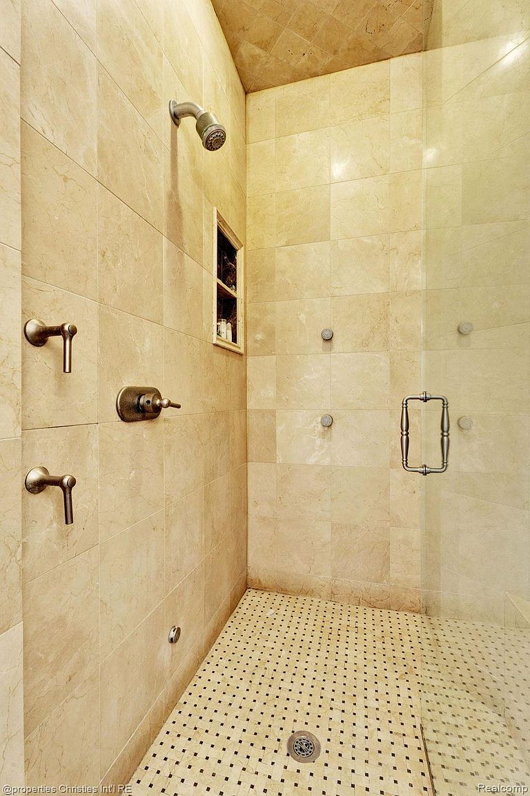 This is a well-lit primary bathroom featuring beige marble tiling on the walls and a mosaic-tiled shower floor. The shower includes a glass door, multiple shower heads, and bronze fixtures. A small niche is built into the wall for storage, creating a luxurious and functional space.