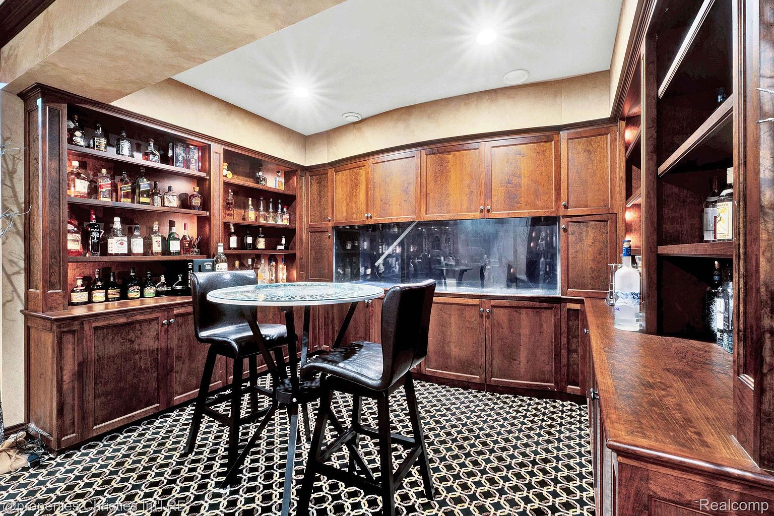This image showcases a sophisticated wine cellar featuring rich, dark wood cabinetry and shelving filled with various bottles. A round table with bar stools sits in the center, creating a cozy tasting area. The patterned floor adds a touch of elegance, while the overall ambiance suggests a luxurious and inviting space for wine enthusiasts.