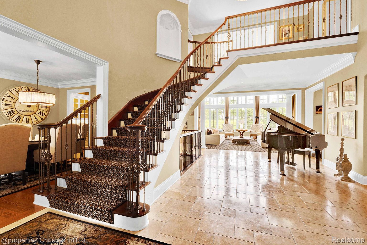 This grand foyer showcases a sweeping staircase with ornate iron railings and patterned carpet. The space is bathed in natural light, highlighting the tile flooring and the elegant piano in the adjacent living area. The overall impression is one of luxury and spaciousness.