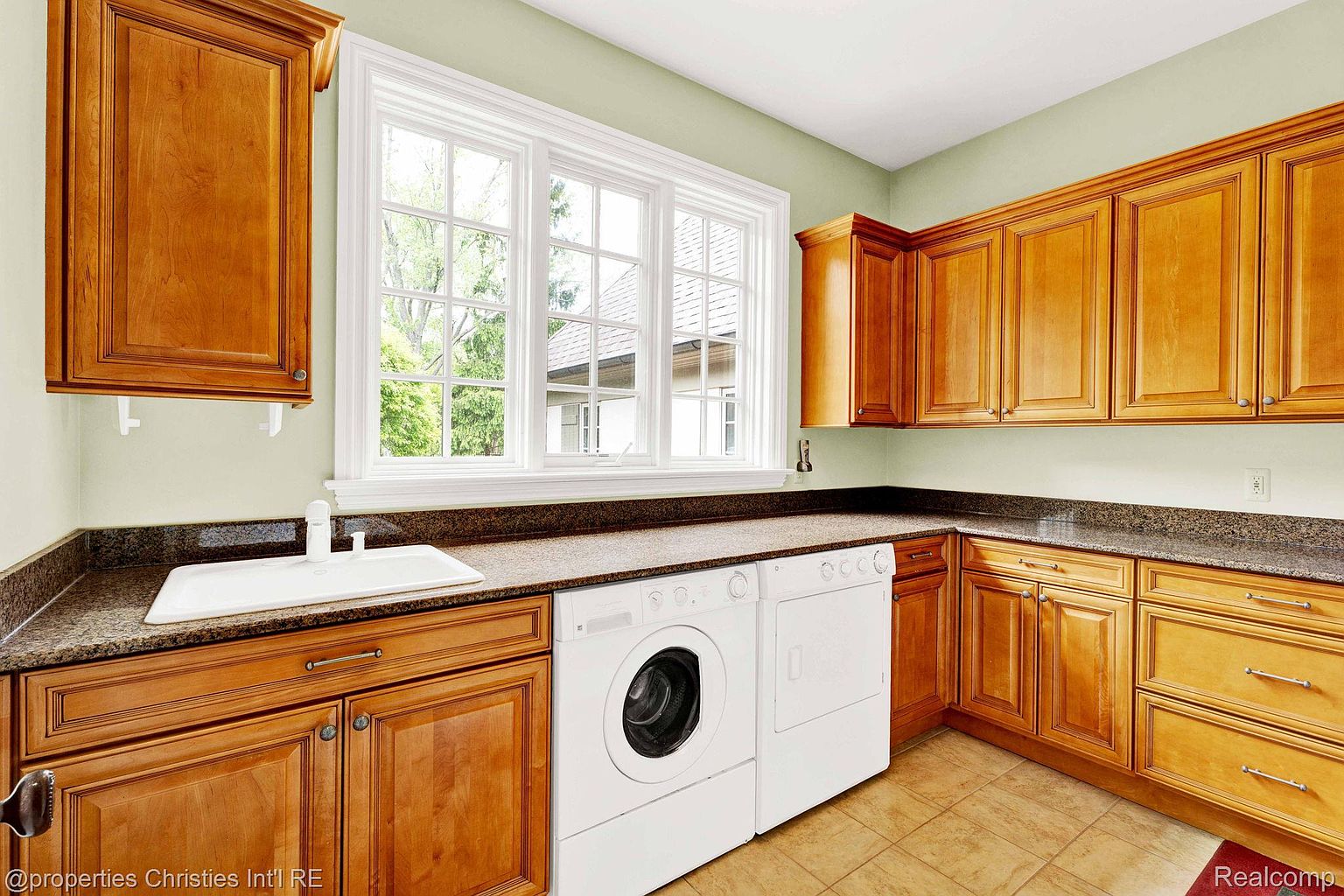 This is a well-lit laundry room featuring wooden cabinetry, granite countertops, and a white sink. A front-loading washing machine and dryer are neatly installed under the counter. The room has a clean and functional design, perfect for a modern home.