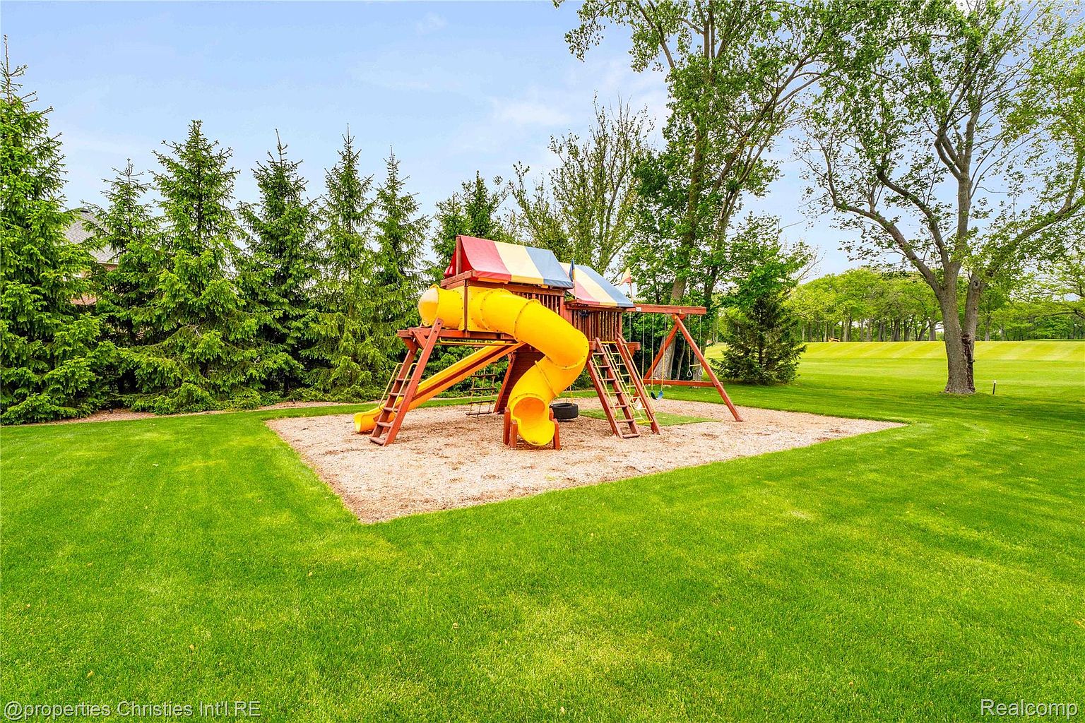 The image showcases a well-maintained yard featuring a colorful playground set. The playground includes a yellow slide, swings, and climbing structures, set on a bed of mulch. Lush green grass surrounds the playground, with mature trees and landscaping providing a scenic backdrop, suggesting a family-friendly environment.