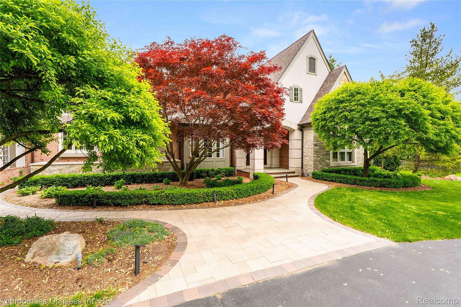 This is a front exterior view of a beautiful home with a well-manicured lawn and landscaping. The house features a combination of stone and stucco exterior, with a prominent red maple tree in the front yard. A curved walkway leads to the front entrance, enhancing the curb appeal of this property.