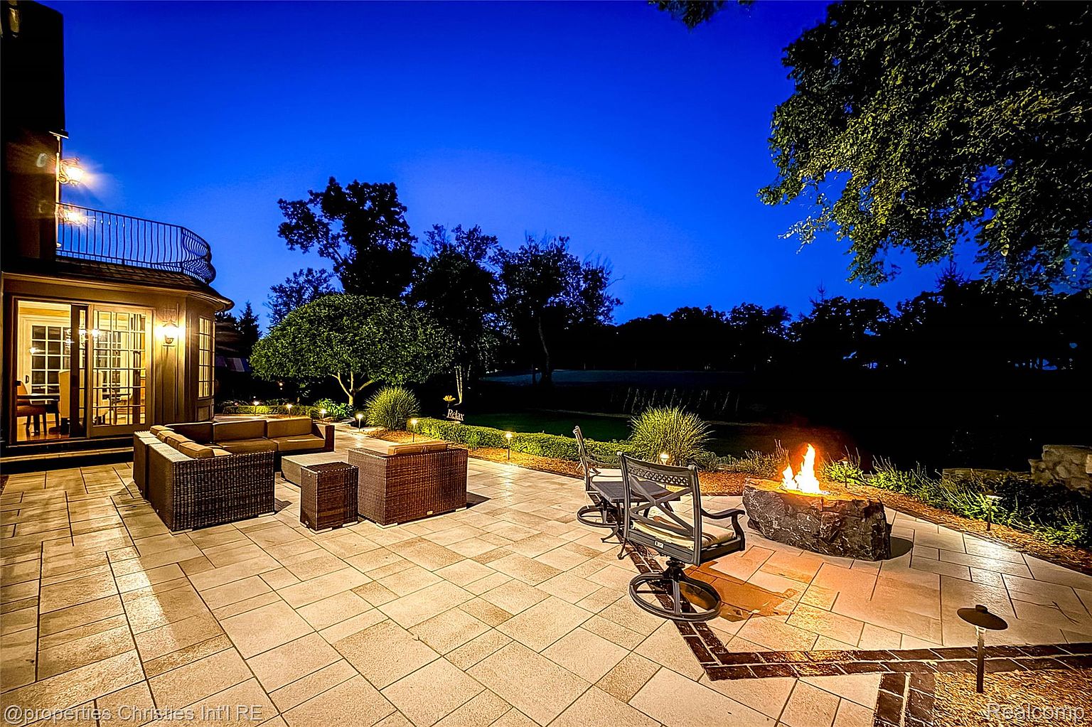 This image showcases a beautifully designed outdoor patio area at dusk, featuring comfortable wicker furniture arranged around a stone fire pit. The patio is paved with large, light-colored stones, and the surrounding landscape includes manicured greenery and mature trees. The warm glow of the fire and the soft lighting create an inviting and luxurious atmosphere, perfect for relaxation and entertaining.
