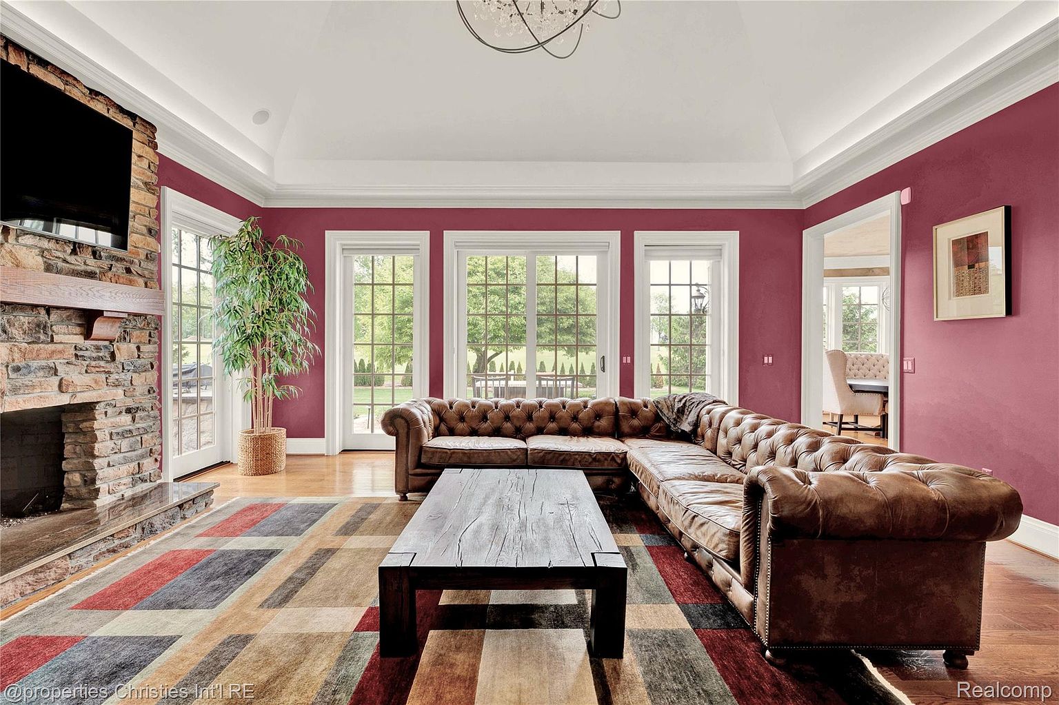 This is a spacious living room featuring a large, tufted leather sectional sofa and a rustic wooden coffee table set on a colorful geometric rug. The room is painted in a deep red hue, complemented by white trim and multiple windows that provide natural light and views of the outdoors. A stone fireplace with a mounted television adds a focal point to the room.