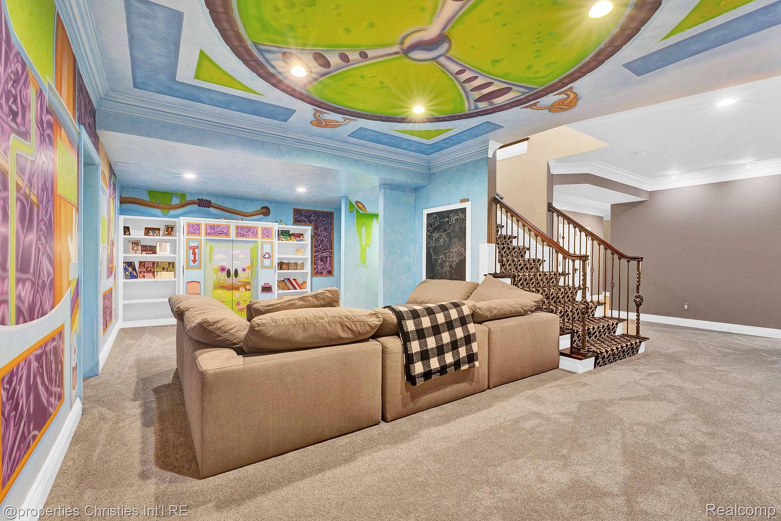 This is a whimsical basement space designed with a playful theme, featuring custom ceiling artwork and colorful wall murals. A large sectional sofa provides ample seating, and built-in shelving offers storage and display space. The staircase leads to the upper level, and the overall impression is a fun and inviting recreational area.