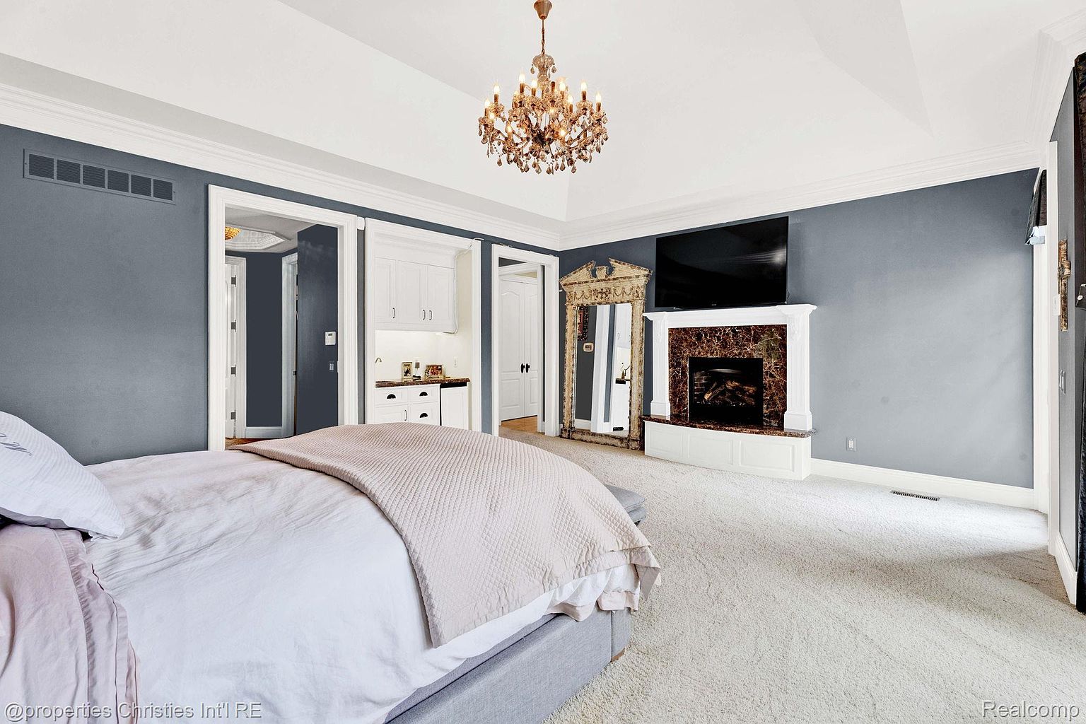 This is a primary bedroom featuring a king-size bed with a light-colored comforter and throw blanket. The room has gray walls, white trim, and a decorative chandelier. A fireplace with a marble surround and a large mirror add to the room's luxurious feel. The perspective is from the foot of the bed, looking towards the fireplace and other room features.