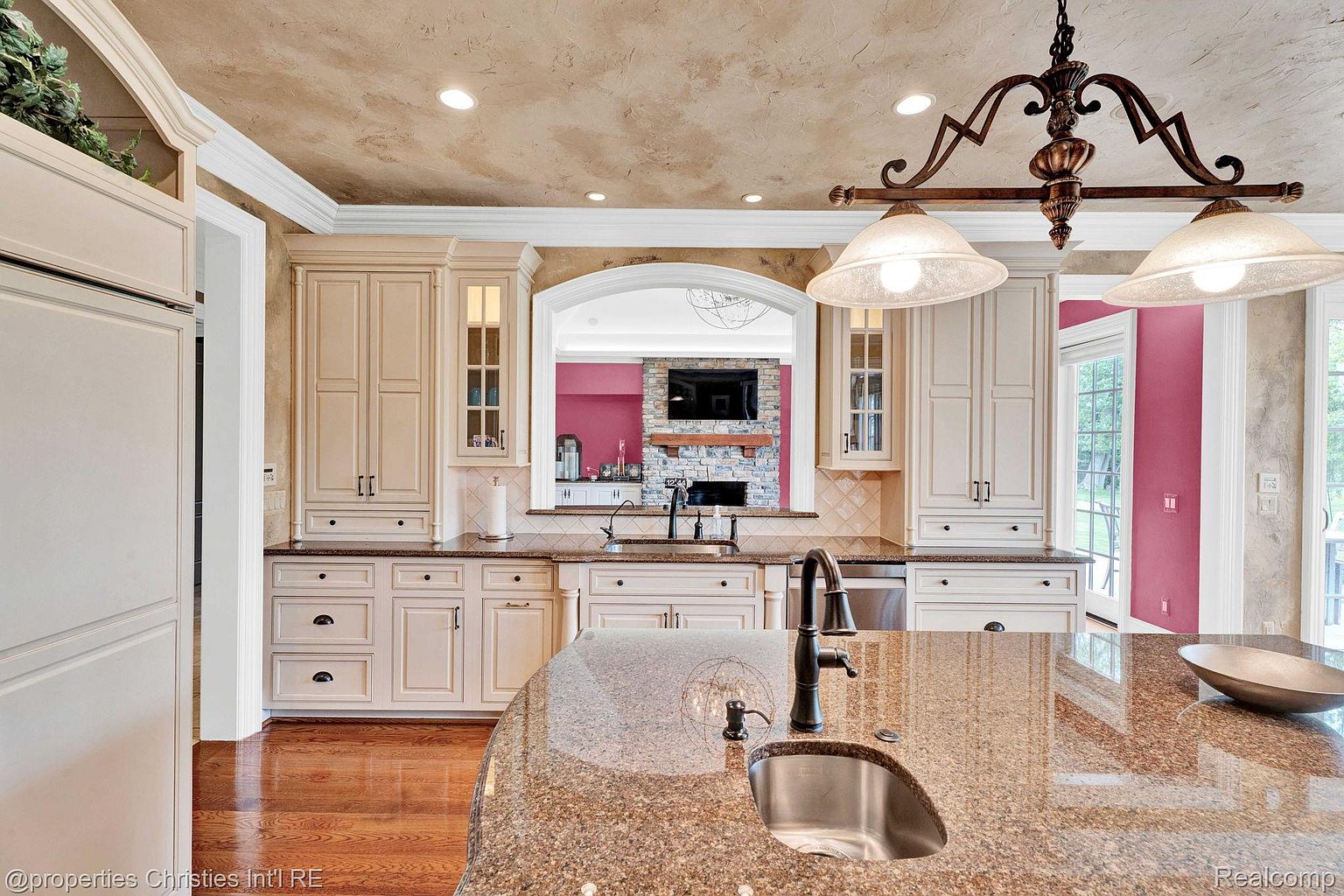This is a well-appointed kitchen featuring custom cabinetry in a light cream color, granite countertops, and a large island with a sink. The kitchen is open to another room through an arched opening, and a decorative chandelier hangs above the island. The overall impression is one of luxury and functionality, perfect for both cooking and entertaining.