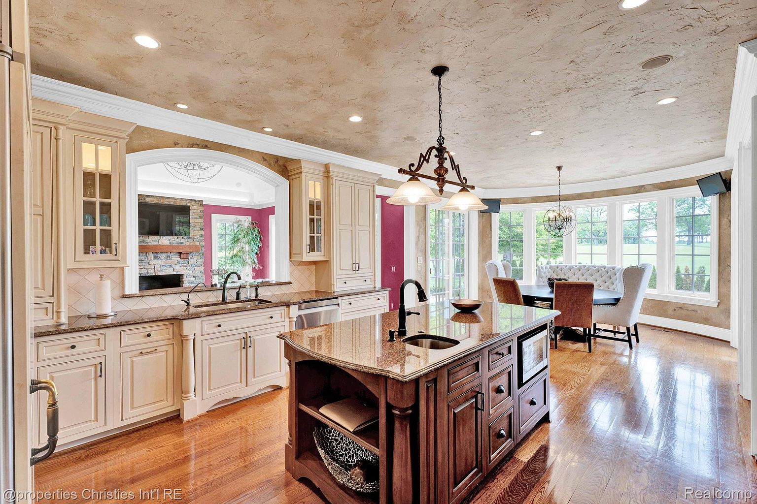 This is a well-appointed kitchen featuring a large center island with a sink and dark wood cabinetry, complemented by lighter-colored perimeter cabinets and granite countertops. The kitchen opens to a dining area with a curved window wall, providing ample natural light. The overall design exudes a sense of luxury and functionality, ideal for both cooking and entertaining.