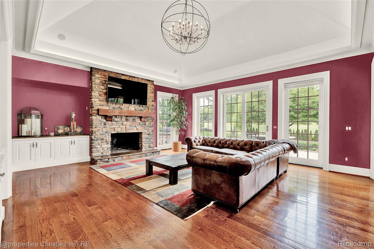 This is an interior shot of a living room featuring a stone fireplace with a mounted television above it. A large, tufted leather sofa sits on a patterned rug in front of the fireplace, and a dark wood coffee table is centered in the space. The walls are painted a deep red, and the hardwood floors are polished, reflecting light from the windows and a decorative chandelier.