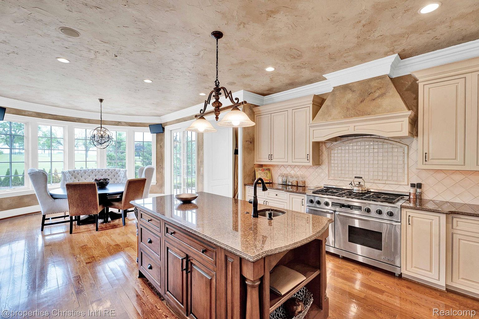 This is a well-appointed kitchen featuring a large center island with a sink and storage, granite countertops, and custom cabinetry. A professional-grade stainless steel range and hood are visible, along with a dining area with a curved window wall. The overall impression is one of luxury and functionality, perfect for both cooking and entertaining.