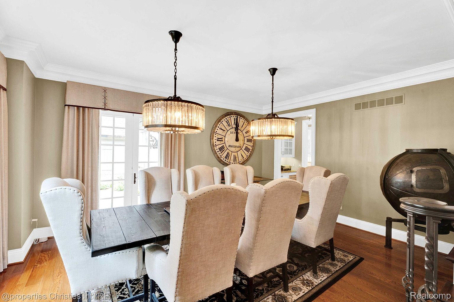 This is an interior shot of a dining room featuring a dark wood table surrounded by upholstered chairs with high backs. Two crystal chandeliers hang above the table, and a large decorative clock adorns the wall. The room is well-lit and has hardwood floors, creating a sophisticated and inviting atmosphere.