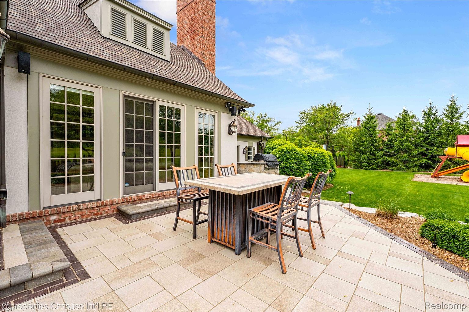 This image showcases a beautifully designed patio area, perfect for outdoor entertaining. The patio features a stone tile surface, a stylish outdoor dining table with chairs, and is adjacent to a well-maintained lawn and garden. The house's exterior is visible, with large windows and a brick accent, adding to the overall appeal of the outdoor living space.