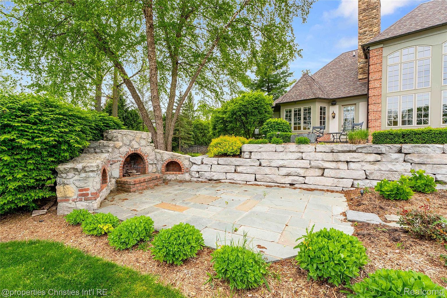 This image showcases a beautifully landscaped backyard featuring a stone patio with a built-in stone fireplace. The patio is surrounded by lush greenery, including manicured bushes and mature trees, creating a serene and private outdoor living space. A stone retaining wall adds architectural interest and defines the garden area adjacent to the house.