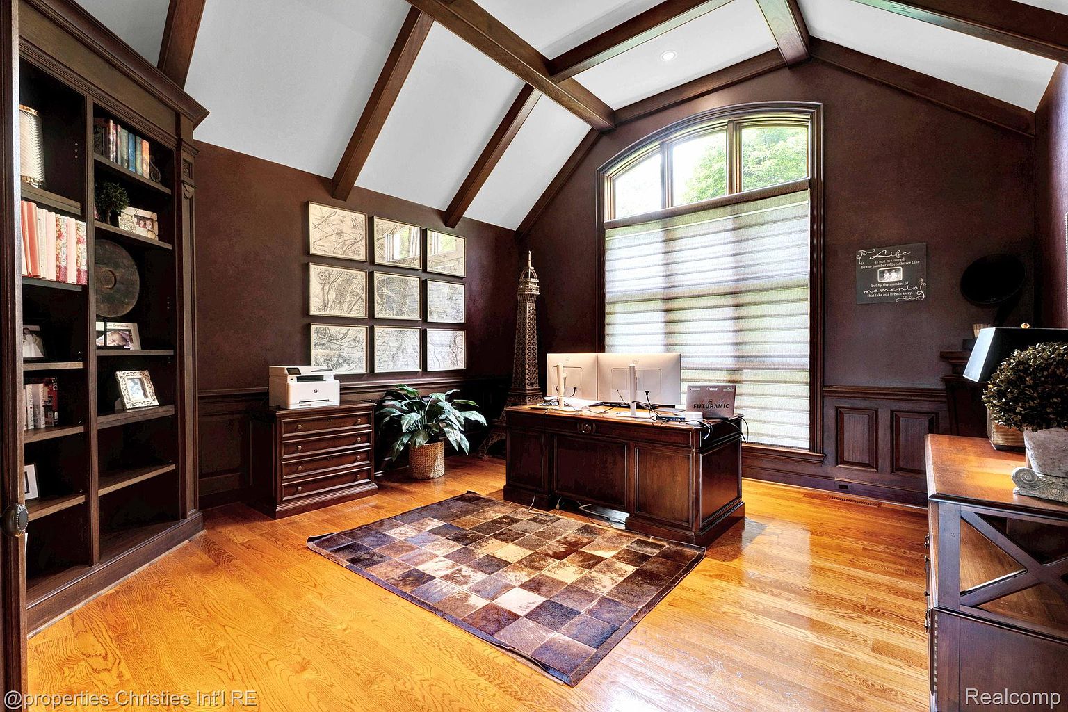 This is a well-appointed home office featuring dark wood finishes, a large desk with multiple monitors, and a built-in bookcase. The room has a sophisticated and professional feel, enhanced by the natural light from the window and the decorative elements like the maps and Eiffel Tower replica. A patterned rug adds warmth to the hardwood floors.