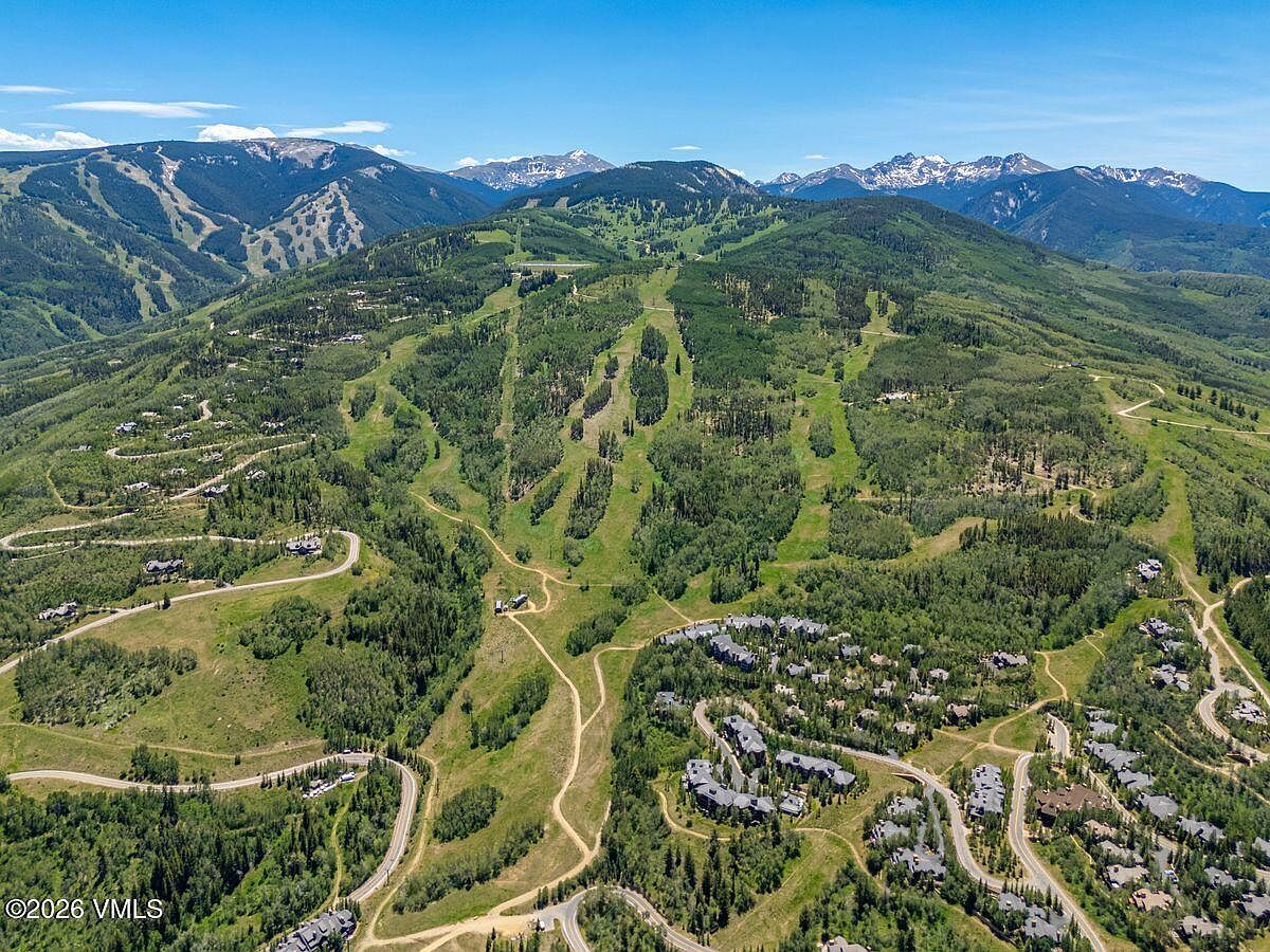 This expansive aerial view captures a sprawling mountain landscape featuring a ski resort nestled within a lush, forested valley. The scene showcases winding access roads, clusters of residential properties, and cleared ski runs that ascend toward the rugged, snow-capped peaks in the distance. The perspective provides a comprehensive look at the community's integration with the natural terrain, emphasizing the serene and scenic environment.