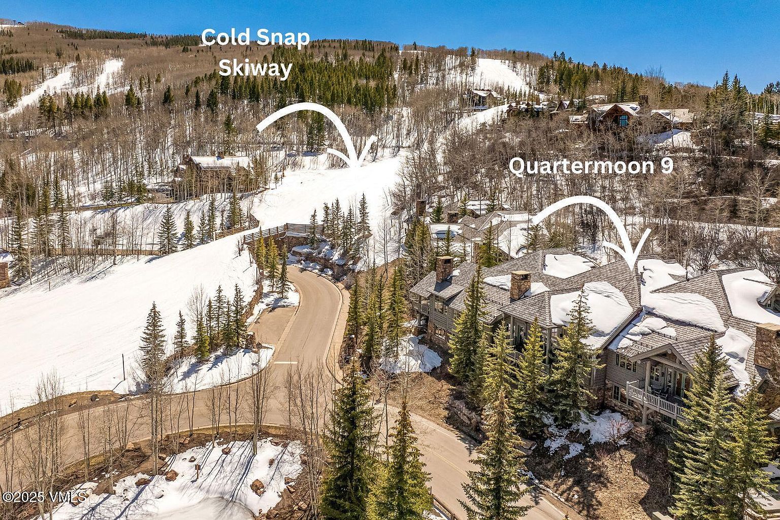 This high-angle aerial view captures a luxury mountain residence nestled within a snowy, forested landscape. The property features a multi-gabled roofline with stone and wood siding, situated directly adjacent to a ski run labeled as Cold Snap Skiway. The scene conveys a serene, upscale alpine lifestyle with a winding road leading toward the home and surrounding evergreen trees dusted with snow.
