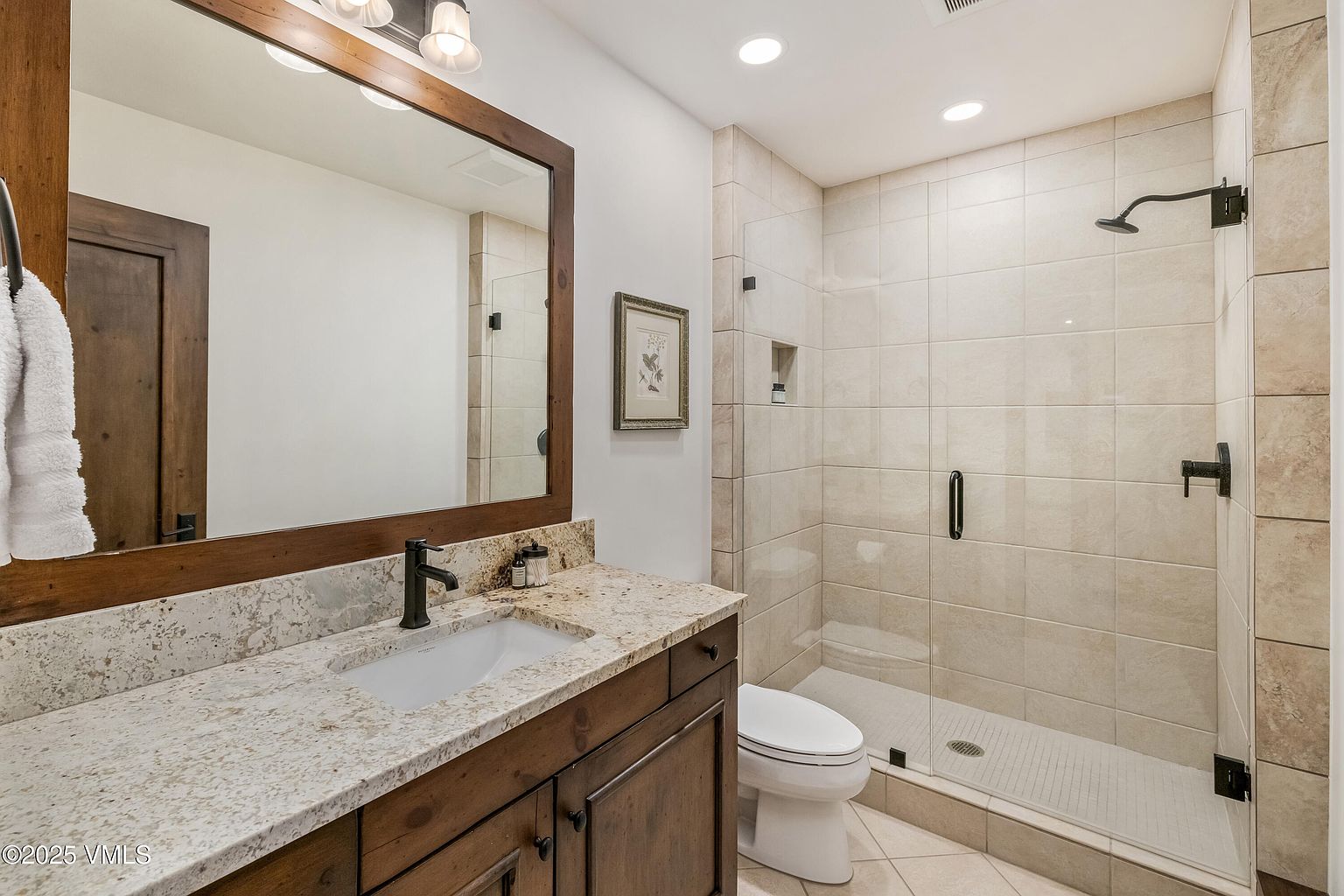 This bathroom features a rustic-inspired vanity with a stone countertop and a dark-toned wood cabinet, complemented by a large mirror framed in matching wood. To the right, a walk-in shower is enclosed by a glass panel and finished with neutral-toned wall tiles. The space is completed by a white toilet and recessed ceiling lighting, creating a clean and functional aesthetic.