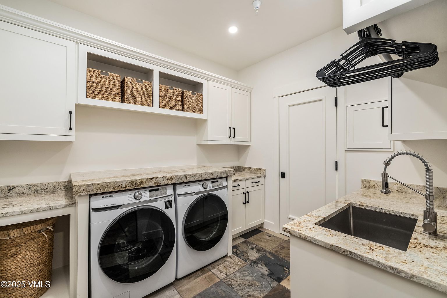 This functional laundry room features white cabinetry, stone countertops, and a stainless steel sink with a gooseneck faucet. A pair of side-by-side Samsung appliances are set beneath the counter, complemented by open shelving with woven storage baskets and a wall-mounted drying rack. The space is finished with a multi-toned, textured tile floor, creating a clean and organized utility area.