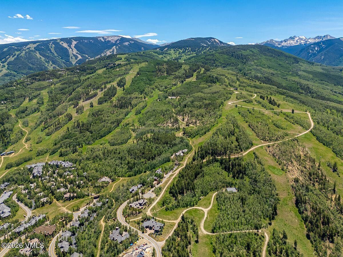 This high-altitude aerial perspective captures a sprawling mountain landscape featuring a residential community nestled among dense forests and ski slopes. The scene highlights the integration of luxury homes into the natural terrain, with winding roads and trails weaving through the lush, green vegetation. In the background, majestic mountain peaks under a clear blue sky provide a dramatic and scenic backdrop for this expansive property setting.