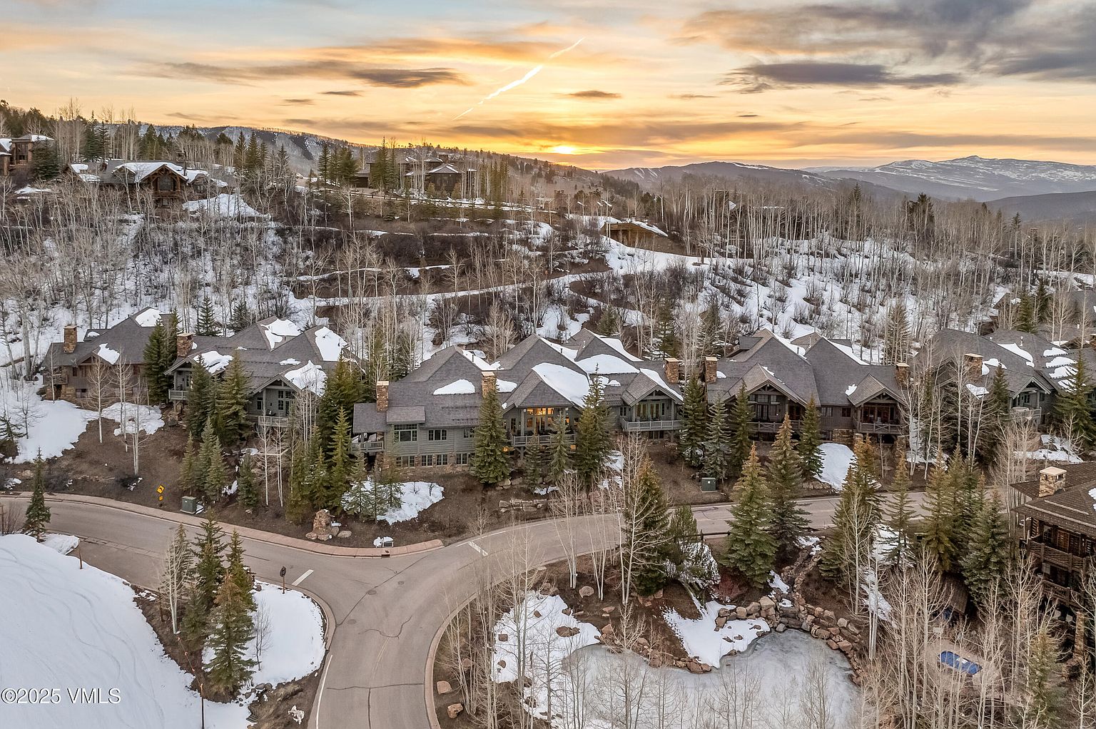 This high-angle aerial perspective captures a cluster of luxury mountain homes nestled into a snowy, forested hillside during a vibrant sunset. The architecture features dark wood siding, complex gabled rooflines, and expansive decks that overlook a winding paved road and a small, partially frozen pond. The scene conveys a serene, upscale alpine lifestyle with the golden light of the setting sun casting long shadows across the rugged, tree-covered landscape.