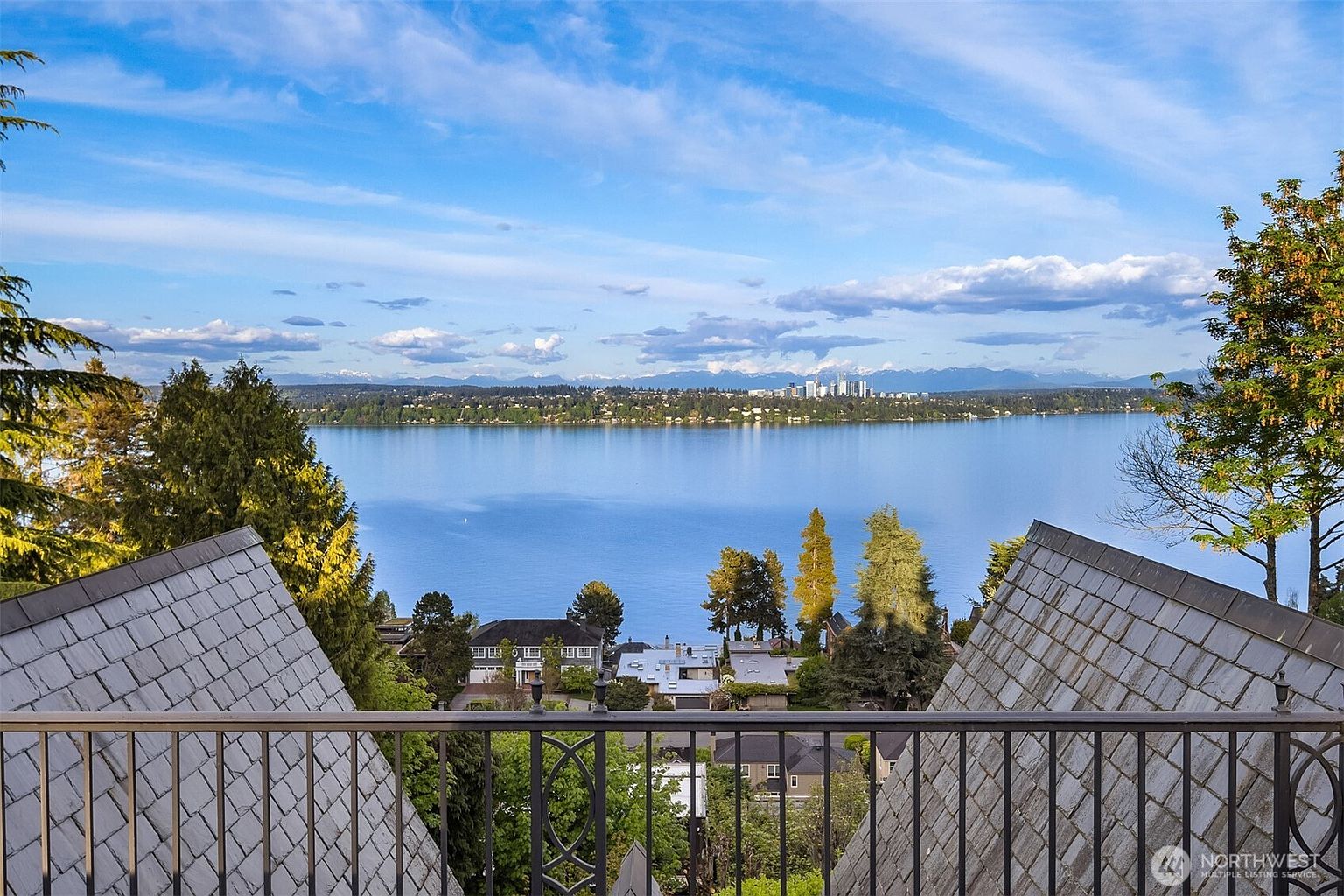 This image captures a breathtaking, elevated view from a balcony overlooking a serene lake and a distant city skyline under a bright, partly cloudy sky. The foreground features a decorative metal railing and the sloped, shingled rooflines of the home, framing the expansive water view. The perspective creates a sense of luxury and tranquility, highlighting the property's prime waterfront location.