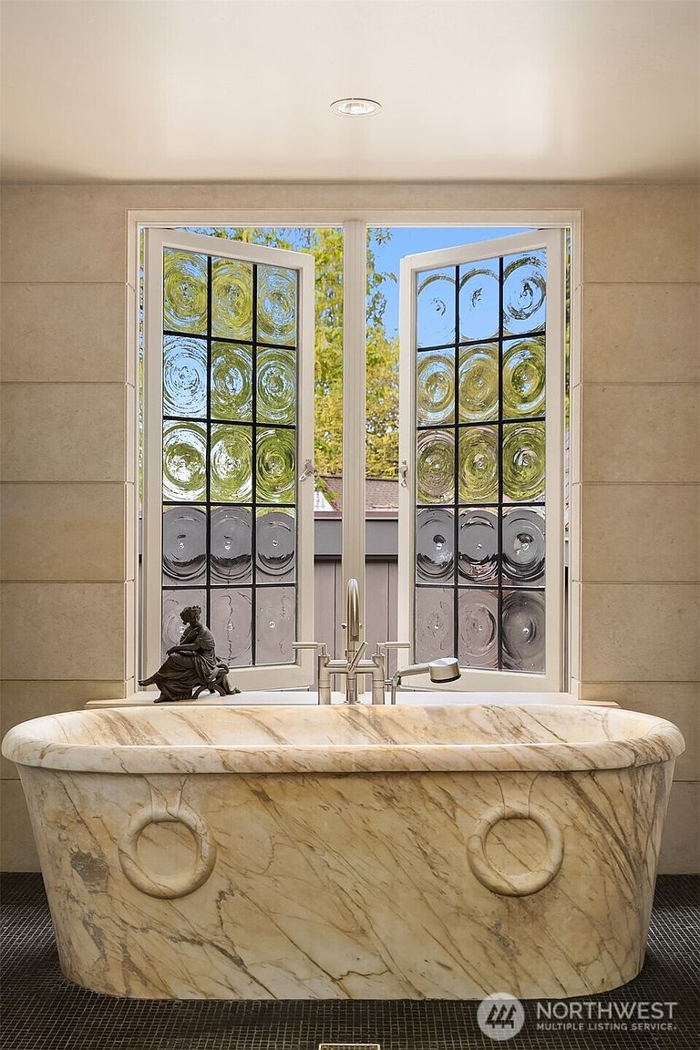 This luxurious primary bathroom features a stunning, hand-carved marble soaking tub as its centerpiece, positioned directly beneath a large, multi-paned window with unique circular glass inserts. The walls are clad in elegant, neutral-toned stone tiles, creating a sophisticated and serene spa-like atmosphere. A classic bronze figurine sits on the window ledge, adding a touch of artistic refinement to the space.