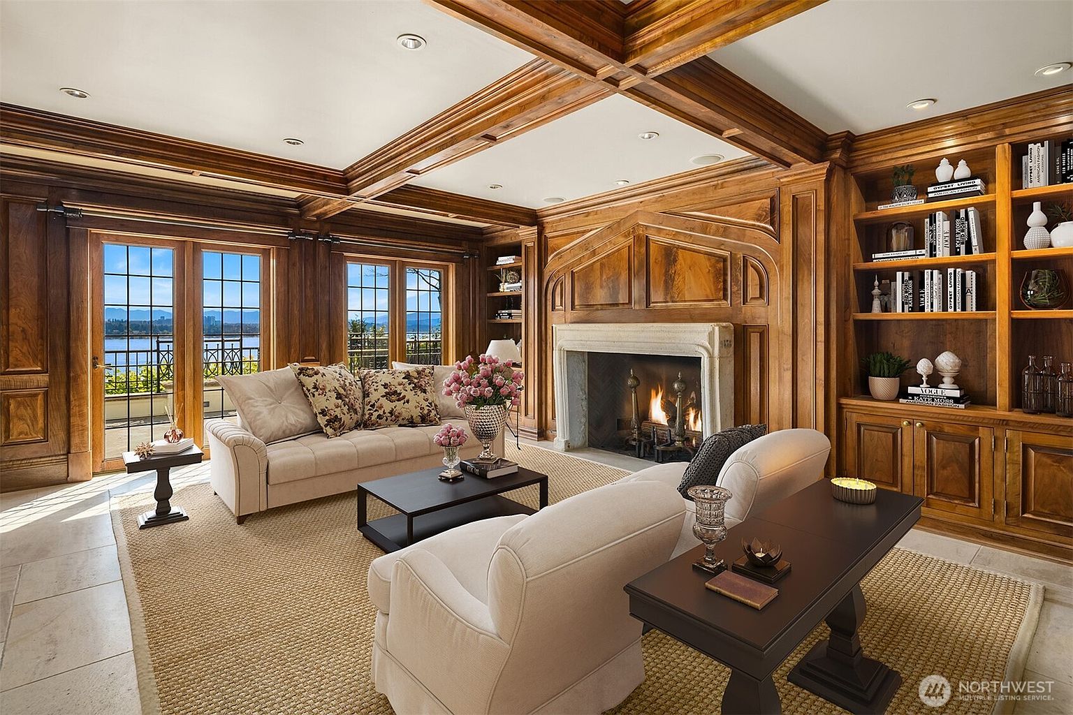 This elegant living room features rich wood-paneled walls, a coffered ceiling, and a grand stone fireplace as the focal point. The space is furnished with two plush, light-colored sofas, a dark coffee table, and built-in bookshelves, all arranged on a textured area rug. Large glass doors open to a balcony, providing a scenic view of the water and mountains beyond, creating a sophisticated and inviting atmosphere.