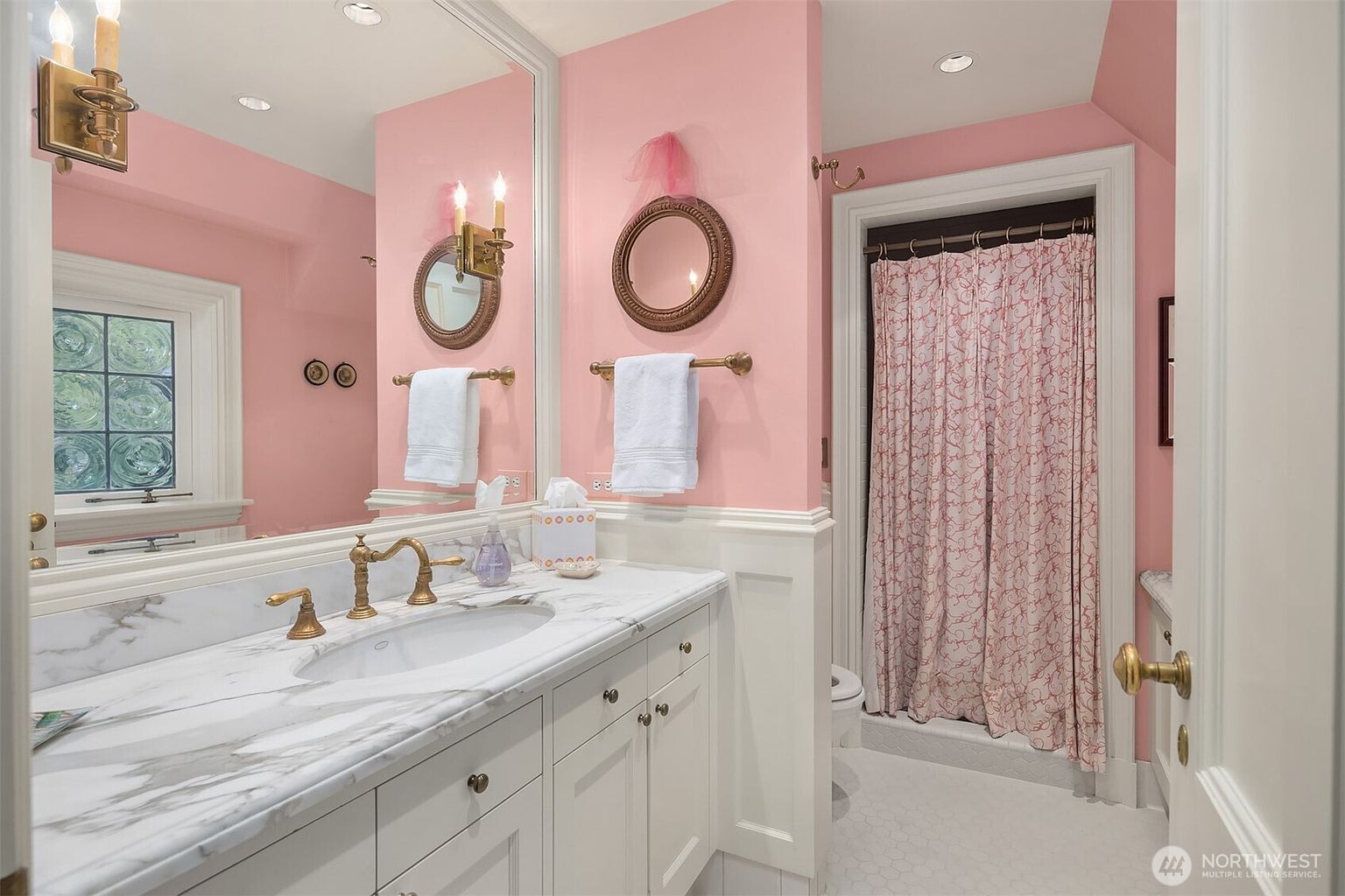This charming bathroom features a vibrant pink wall color contrasted by crisp white wainscoting and a marble-topped vanity. The space is accented with elegant gold-toned fixtures, including wall-mounted sconces and a circular mirror, creating a whimsical and polished aesthetic. A shower area with a patterned curtain is tucked into the corner, completing this well-appointed and bright guest bathroom.