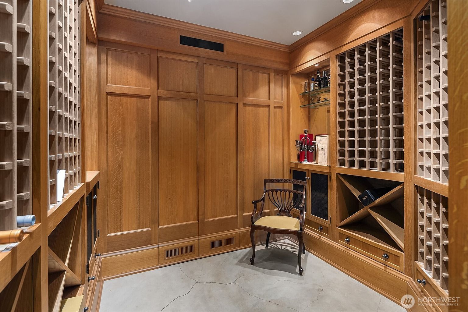 This sophisticated wine cellar features extensive custom oak cabinetry with built-in wine racks and a dedicated display area. A classic wooden chair sits in the center of the room, which is finished with a smooth concrete floor and elegant paneling. The space exudes a refined, traditional atmosphere, perfect for wine storage and quiet appreciation.