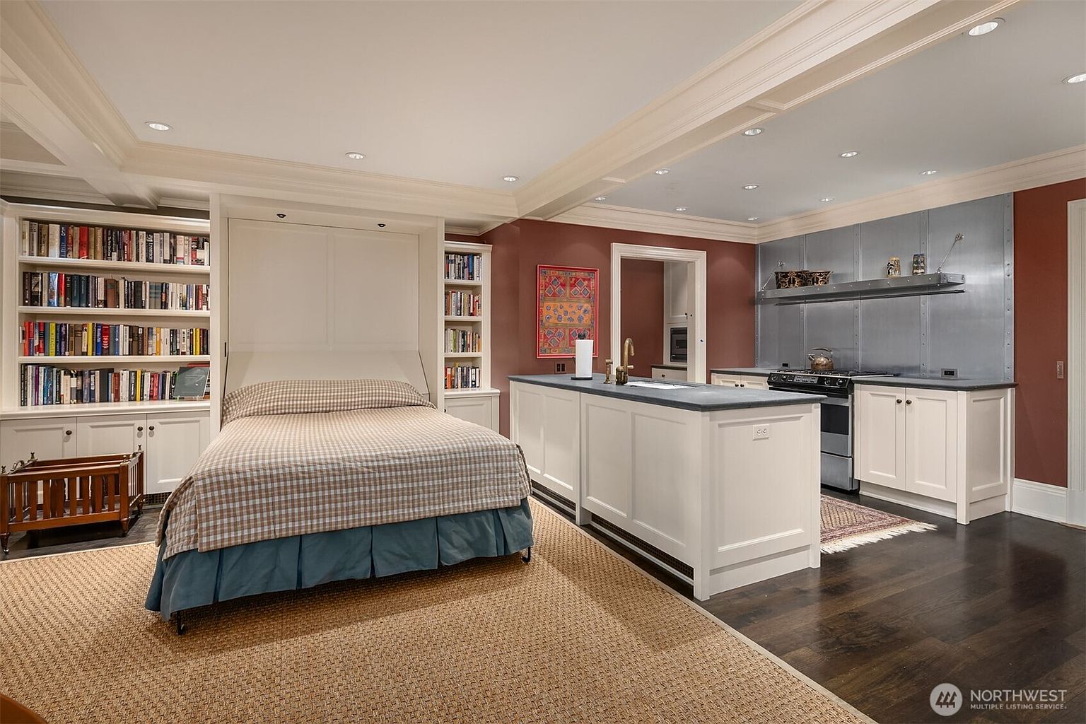 This versatile studio-style room features a space-saving Murphy bed integrated into a wall of custom white bookshelves, creating a seamless blend of sleeping and living areas. To the right, a functional kitchen area with a central island, stainless steel backsplash, and white cabinetry provides modern convenience. The room is anchored by dark hardwood floors and a large textured area rug, offering a sophisticated and multi-purpose living environment.