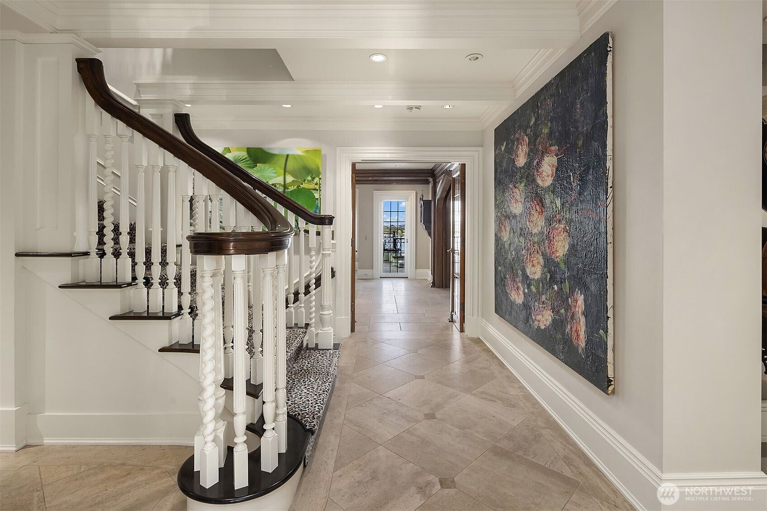 This elegant foyer features a grand staircase with a dark wood handrail and white balusters, accented by a striking animal-print runner. A large, moody floral painting hangs on the right wall, contrasting beautifully with the neutral, light-colored tile flooring. The perspective looks down a central hallway toward a bright doorway, creating a sense of depth and sophisticated architectural flow.