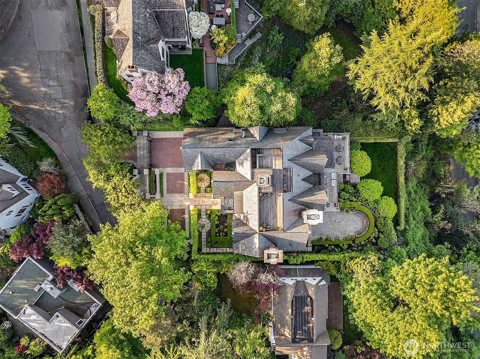 This high-angle aerial view captures a sprawling, multi-gabled estate nestled within a lush, mature landscape. The property features intricate stone pathways, a formal garden, and a spacious rear patio, all surrounded by dense, vibrant greenery. The perspective highlights the architectural complexity of the roofline and the seamless integration of the home into its private, wooded environment.